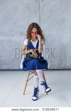 Adorable Little Girl In Dress, Sitting On Chair And Looking Through Eyeglasses Over Concrete Backgro