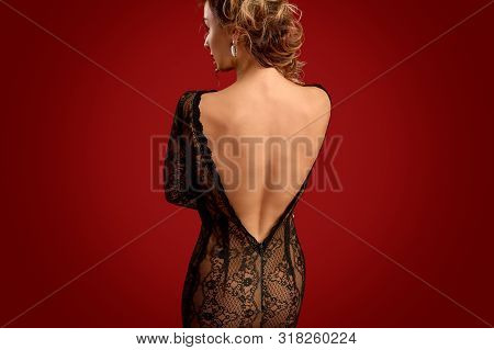 Back View Of Sensual Female In Backless Lace Dress Looking Away While Standing Against Bright Red Ba