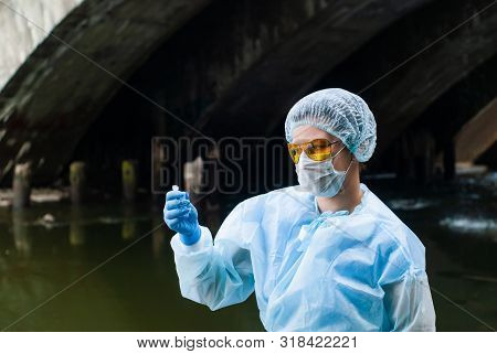 Female Ecologist Or Epidemiologist Takes Test-tube Water Analysis In A City River Under An Old Bridg