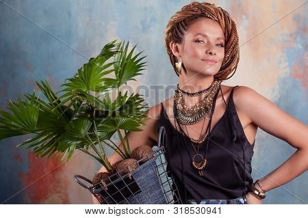 Southern Flavor. A Young Tanned Woman With Afro Braids Smiles Holding A Big Pot With A Tropical Plan