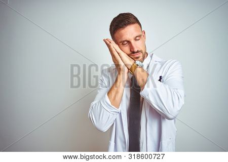 Young handsome doctor man wearing white profressional coat over isolated background sleeping tired dreaming and posing with hands together while smiling with closed eyes.