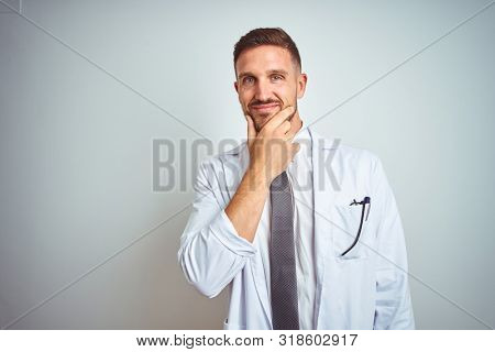 Young handsome doctor man wearing white profressional coat over isolated background looking confident at the camera smiling with crossed arms and hand raised on chin. Thinking positive.