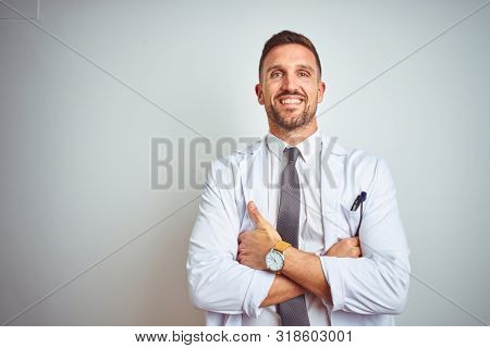 Young handsome doctor man wearing white profressional coat over isolated background happy face smiling with crossed arms looking at the camera. Positive person.
