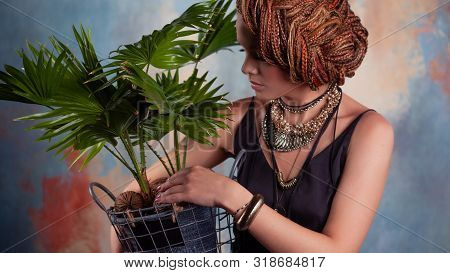 Southern Flavor. A Young Tanned Woman With Afro Braids Smiles Holding A Big Pot With A Tropical Plan
