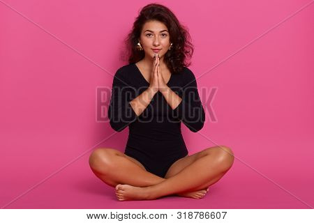Studio Shot Of Young Woman Wearing Kombidress, Sitting On Floor In Lotus Pose, Adorable Girl Holding