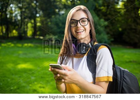 Portrait Of Beautiful Student Girl With Daypack Using Her Smartphone On The Park Lawn