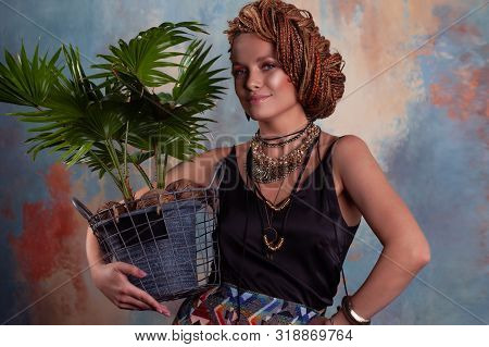 Southern Flavor. A Young Tanned Woman With Afro Braids Smiles Holding A Big Pot With A Tropical Plan