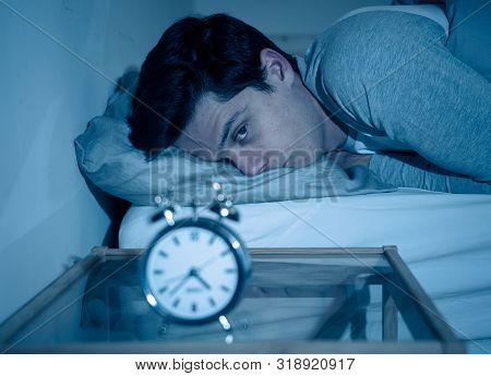 Portrait Of Young Man In Bed Staring At Alarm Clock Trying To Sleep Feeling Stressed And Sleepless