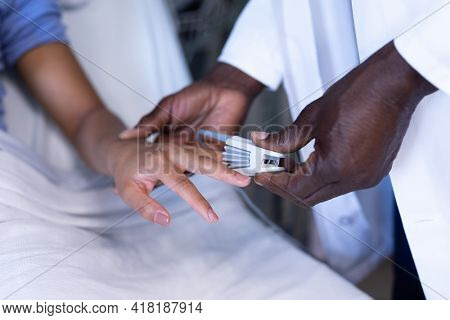 Midsection of african american male doctor putting oximeter on finger of woman in hospital bed. medicine, health and healthcare services.
