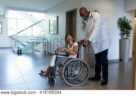 Smiling african american male doctor pushing mixed race girl in wheelchair through hospital corridor. medicine, health and healthcare services.