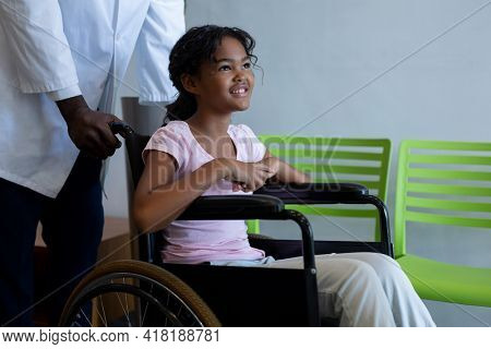 African american male doctor pushing smiling mixed race girl in wheelchair through hospital corridor. medicine, health and healthcare services.