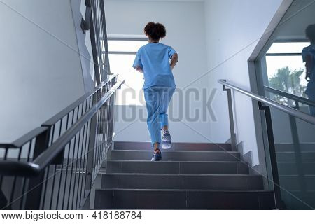 Rear view of mixed race female doctor wearing scrubs running up stairs in hospital. medical professional at work.