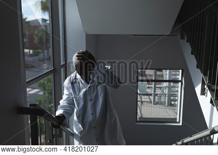 African american male doctor going upstairs on hospital staircase holding head and worrying. medicine, health and healthcare services.