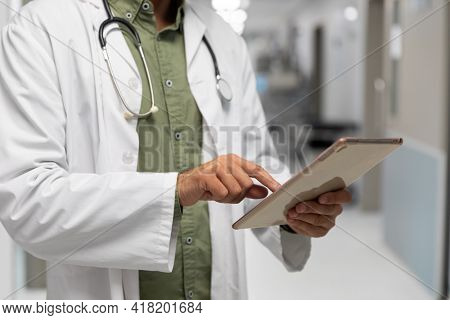 Midsection of caucasian male doctor standing in hospital corridor looking at tablet. medicine, health and healthcare services.