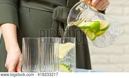 Drink Water. Woman's Hand Pouring Fresh Pure Water With Pieces Of Lemon From Pitcher Into Glass In T