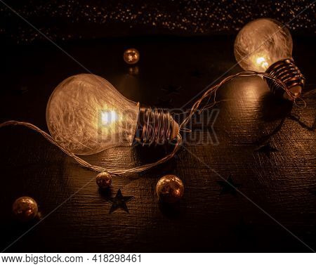 Close Up Of Two Light Bulbs Containing Tiny White Featers On A Dark Surface And Decorated With Glitt