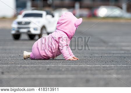 A Little Child In A Pink Dress Crawling On The Asphalt In A Car Parking Lot