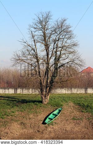 Light Green With Dark Brown Edge Elongated Wooden River Boat Left On Grass And Dry Soil Covered Rive