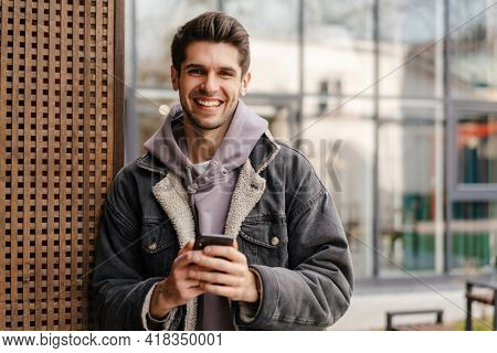 Smiling young white man in casual cothes using mobile phone outdoors in the city, leaning on a wall