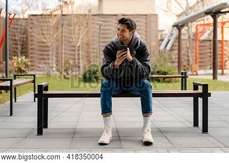 Smiling young white man in casual cothes using mobile phone outdoors in the city, sitting on a bench