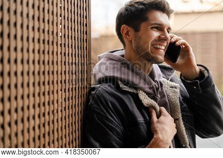 Smiling young white man in casual cothes using mobile phone outdoors in the city, leaning on a wall, talking