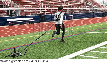 A High School Track Runner Is Pulling A Sled With Weights On It Across A Green Turf Field During Str