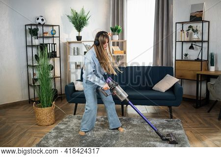 Charming Young Woman In Modern Headphones Enjoying Favorite Music While Cleaning House With Hand Hel