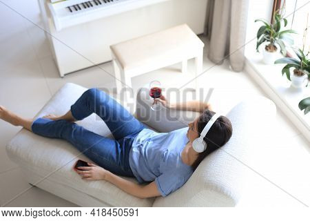 Beautiful Brunette Woman In Earphones Sitting On Sofa Listening To Music With Closed Eyes, Drinking