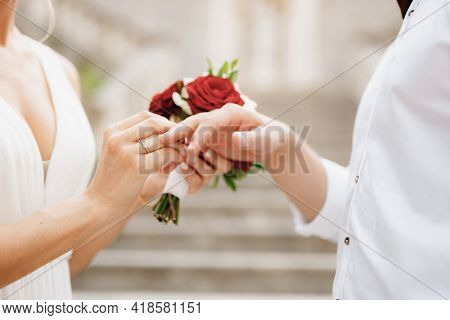 The Bride Puts A Wedding Ring On The Grooms Finger And Holds A Bouquet Of Red And White Roses In Her