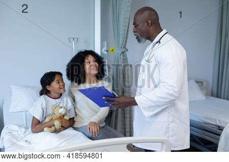 African american male doctor talking to mixed race mother and her sick daughter, in hospital bed. medicine, health and healthcare services.