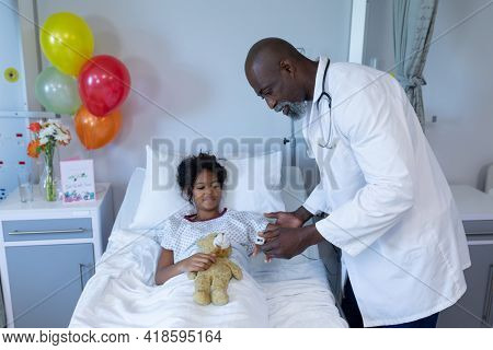 African american male doctor putting pulse oximeter on sick mixed race girl in hospital bed. medicine, health and healthcare services.