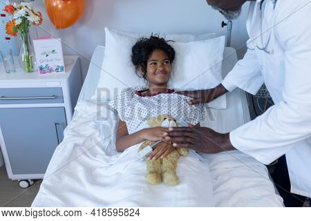 African american male doctor reassuring smiling sick mixed race girl in hospital bed holding teddy. medicine, health and healthcare services.