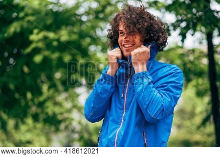 Smiling Man With Curly Hair Wearing Blue Raincoat During Rain Outside. Positive Male In Blue Raincoa