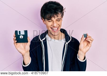 Handsome hipster young man holding floppy disk and sdxc card smiling and laughing hard out loud because funny crazy joke. 