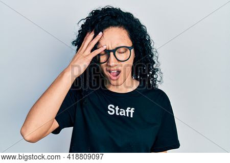 Young hispanic woman with curly hair wearing staff t shirt yawning tired covering half face, eye and mouth with hand. face hurts in pain. 
