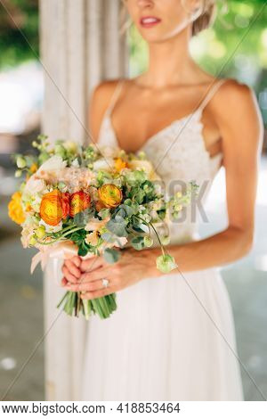 The Bride Stands Among White Columns Braided With Green Vines And Holds A Wedding Bouquet With Orang