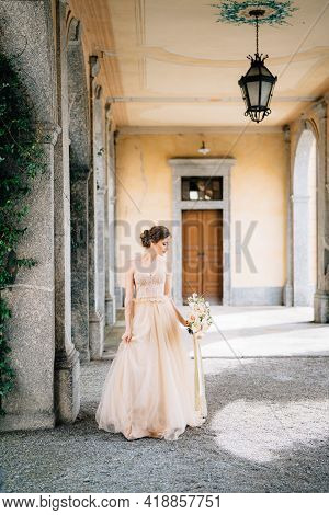 Bride In A Beautiful Dress With A Bouquet Of Pink Flowers Stands In An Arched Corridor Near The Gree