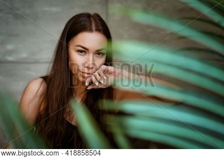 Beautiful Young Woman Closeup Portrait Through Green Leaves Of A Flower On A Gray Wall Background