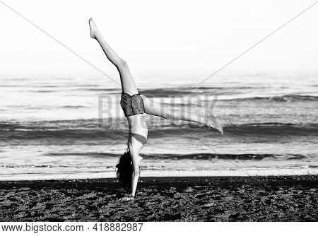 Athletic Young Girl Performs Gymnastic Exercises By Doing The Wheel On The Beach By The Sea With Bla