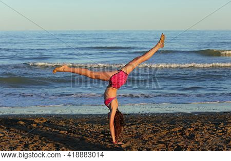 Athletic Young Girl In Swimsuit Performs Gymnastic Exercises On The Beach By The Sea
