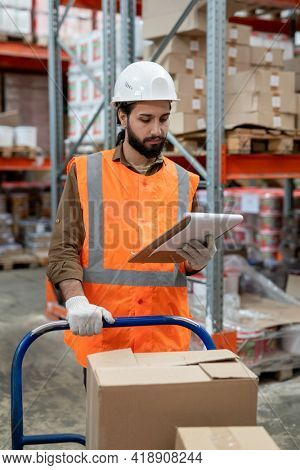 Serious busy young mixed race warehouse worker in hardhat reading consignment note while examining item placement