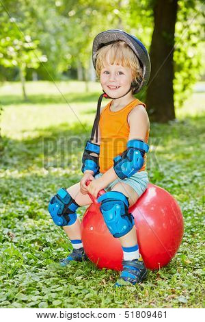 Smiling little boy in protective equipment sits on red ball for jumping on lawn in park