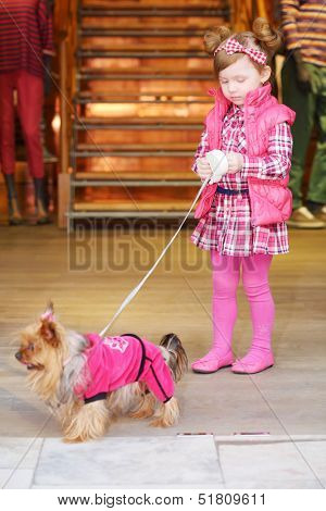 Little cute girl in pink dress leads small dog on leash in clothes in store.