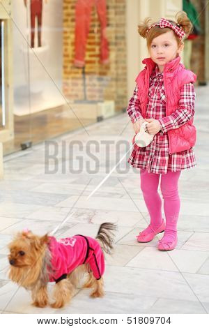 Little beautiful girl in dress leads small dog on leash in clothes in store.