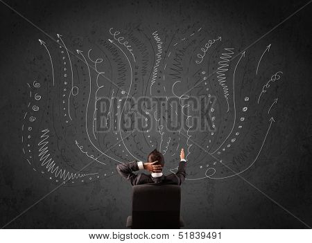 Thoughtful young businessman sitting and deciding in front of a chalkboard with sketched arrows and lines in different directions