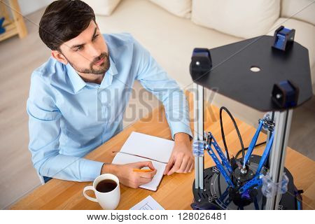 Everything under control. Pleasant delighted bearded man sitting at the table and making notes while using 3d printer