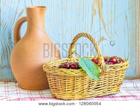 Wicker basket with ripe red cherries and a clay jug on a checkered tablecloth on wooden painted blue background