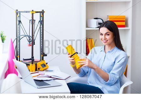 Full of positive emotions.  Pleasant beautiful smiling delighted woman sitting at the table and holding model printed on the 3dprinter while expressing gladness