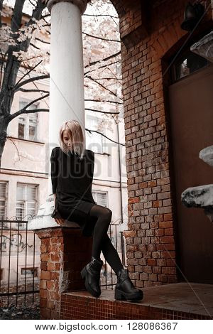A girl with blonde hair has leaned the elbows on a column from a brick and looks down having bent a knee.