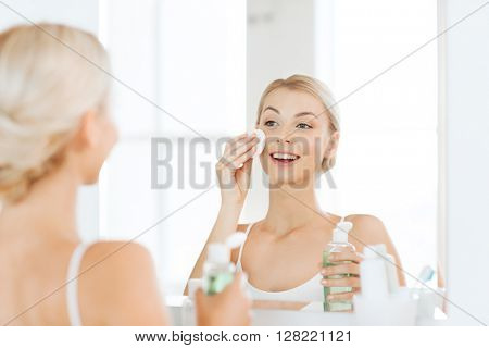 beauty, skin care and people concept - smiling young woman applying lotion to cotton disc for washing her face at bathroom